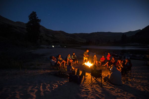 a group of people sit around a glowing red campfire at dusk